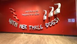 A bold red wall features the text “…and she lived happily ever after” in white serif letters, with the phrase “WITH HER THREE DOGS!” in playful blue block letters below, framed by three wavy hair sculptures on the left and three white dog tails emerging from the wall on the right.