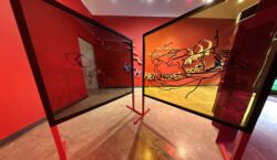 The plexiglass panels, arranged in a zigzag, display black line drawings of baby Sora interacting with her mother, creating layered visuals with the red gallery walls and text in the background.