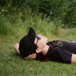 Naomi Watkins laying on their back in the grass with a black shirt, hat and sunglasses, with foliage in the background.